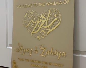 May include: A gold and beige welcome sign for a wedding reception. The sign features Arabic calligraphy in a heart shape, the names "Ayaz & Zahiya", the text "AND WE CREATED YOU IN PAIRS" with the Quran reference "QURAN 78:8", and the date "NOV 03, 2024".