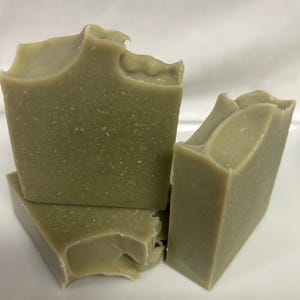 Puede incluir: Cuatro barras rectangulares de jabón artesanal de diferentes tamaños, dispuestas sobre una superficie blanca. El jabón tiene un tono verde claro con motas visibles y una textura ligeramente irregular. Los bordes de las barras de jabón tienen una forma ondulada y orgánica.