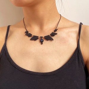 May include: A black macrame necklace with a dark blue gemstone pendant. The necklace features a geometric design with gold beads.