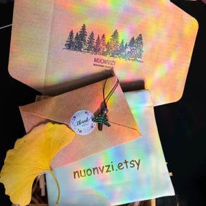 Puede incluir: Un sobre de papel marr&oacute;n con una pegatina de agradecimiento y un peque&ntilde;o amuleto de &aacute;rbol de Navidad de cuentas verdes. El sobre est&aacute; dirigido a nuonvzi.etsy.
