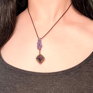 May include: A necklace featuring a dark brown cord, a light purple macrame leaf-shaped accent, and a square dark purple gemstone pendant encased in a decorative brown woven frame that hangs centered from the cord.