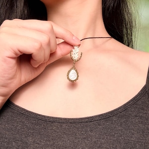 May include: A necklace featuring a teardrop-shaped white stone pendant encased in a textured bronze-colored wire frame. Above the stone, a small white coral-like element is attached to the frame, suspended from a thin dark brown cord.