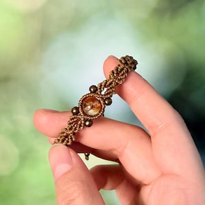May include: A brown macrame bracelet featuring a central round amber-colored bead flanked by two smaller metallic bronze beads, with intricate knotted cord patterns extending along the band to create a textured and detailed wrist accessory.