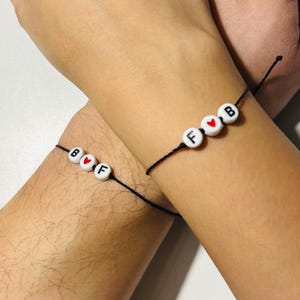 May include: Two black string bracelets with white beads spelling out "BFF" and "FB" with a red heart bead between the letters.