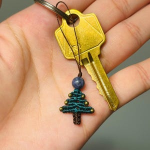 May include: A small, green and brown, crocheted Christmas tree keychain with a blue bead and gold accents. The keychain is attached to a gold key.