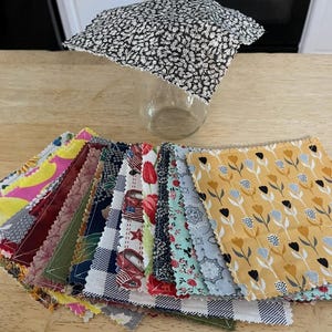May include: A collection of square fabric food covers with pinked edges, featuring various patterns including yellow tulips, red trucks, floral prints, and gingham checks. One cover is draped over a clear glass jar, while others are stacked below.