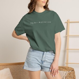 May include: A dark sage green short-sleeve t-shirt featuring white printed text on the chest that reads lim f(x) = R · (I)² · Z · E (1.0). The shirt has a crew neckline and a standard fit, paired with light blue denim shorts with a cuffed hem.