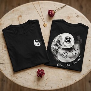 Könnte beinhalten: Zwei schwarze T-Shirts mit Yin-Yang-Grafik. Das linke T-Shirt hat ein kleines Yin-Yang-Symbol auf der Brust. Das rechte T-Shirt zeigt ein großes Yin-Yang-Design mit Berglandschaft, Baum, Fledermäusen und dem Text One, Two, Connect.