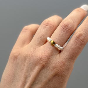 Könnte beinhalten: Ein Perlenring mit einem goldfarbenen herzförmigen Anhänger. Der Ring besteht aus kleinen, weißen Perlen und hat ein kleines, goldenes Herz in der Mitte. Der Ring wird an einem Finger getragen.