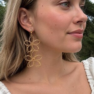 Könnte beinhalten: Ein Paar goldfarbene Ohrhänger mit zwei übereinander angeordneten, offenen Drahtblütenformen. Die Ohrringe sind an einem kleinen, kreisförmigen Verschluss befestigt. Das Design besteht aus dünnen, durchgehenden Drahtschleifen in Blütenform.