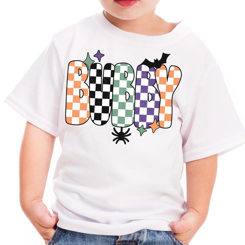 May include: White t-shirt with the word "BUBBY" in orange, black, green, and purple checkered letters. A black bat and spider are above and below the text, with star accents. The shirt is for children.