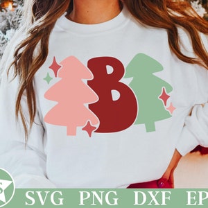 May include: White sweatshirt with a Christmas-themed design. The design features a large burgundy letter "B" flanked by a pink and a green Christmas tree, with star accents. The bottom of the image shows the text "SVG PNG DXF EPS".