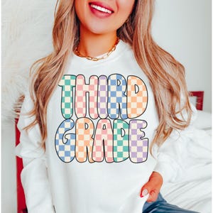 May include: White sweatshirt with the text "THIRD GRADE" in colorful checkered letters.