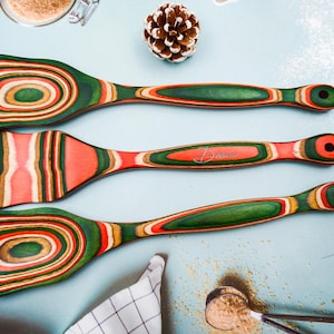 Könnte beinhalten: Drei Küchenutensilien aus Holz mit einem farbenfrohen Streifendesign in Rot, Grün und Gold. Die Utensilien umfassen zwei Löffel und einen Spatel. Die Griffe sind grün und die Enden haben ein Loch zum Aufhängen. Auf dem Spatel steht der Name "Dena".