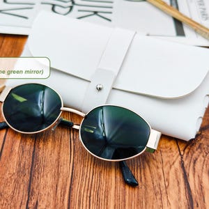 May include: Gold-framed sunglasses with green mirrored lenses rest on a wooden surface next to a white leather case. The sunglasses have a classic round shape and dark temples. The case is closed with a strap.