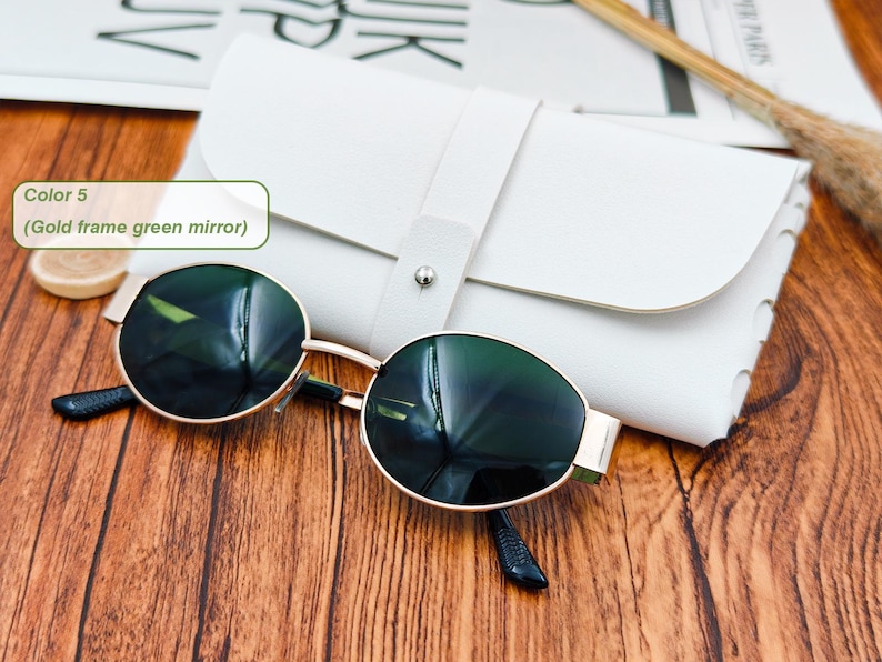May include: Gold-framed sunglasses with green mirrored lenses rest on a wooden surface next to a white leather case. The sunglasses have a classic round shape and dark temples. The case is closed with a strap.