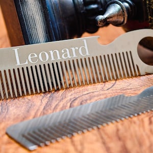 May include: A silver-colored comb with the name "Leonard" engraved on it. The comb has a bottle opener feature and a circular hole. Another comb with a zig-zag edge is also visible. Both are on a wooden surface.