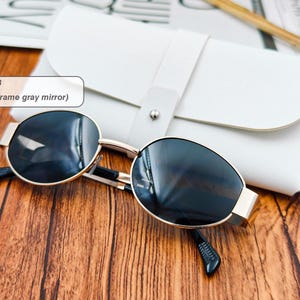 May include: Gold-framed sunglasses with gray mirrored lenses rest on a wooden surface next to a white leather case. The sunglasses have a classic design with a rounded shape. The text in the image says "Color 3 (Gold frame gray mirror)".