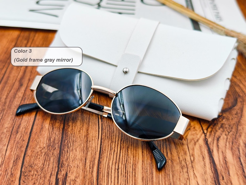 May include: Gold-framed sunglasses with gray mirrored lenses rest on a wooden surface next to a white leather case. The sunglasses have a classic design with a rounded shape. The text in the image says "Color 3 (Gold frame gray mirror)".