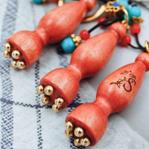 Puede incluir: Primer plano de tres herramientas de masaje de madera roja con puntas de bola doradas. Cada herramienta tiene un diseño decorativo y está ensartada con un cordón negro, cuentas turquesas y rojas y detalles dorados. Sobre una superficie estampada.