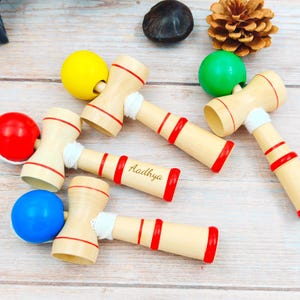 May include: Wooden kendama toys with red accents and colorful balls in yellow, green, red, and blue. One kendama has the name "Aadhya" engraved on it. The toys are arranged on a light wood surface.