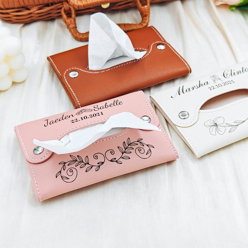 May include: Three leather tissue box covers in brown, pink, and white. The pink cover has a floral design and the names "Jaeden Sabelle" and the date "22.10.2021". The white cover has the names "Marsha Clinton" and the date "22.10.2021".