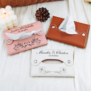 May include: Three personalized tissue box holders in pink, brown, and white. Each holder features a decorative design and the names and date of a wedding. The pink holder has a floral design, while the white one has flowers.