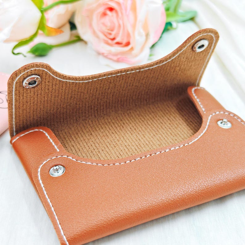 May include: A brown leatherette card holder with white stitching and snap closures. The interior is a textured brown fabric. The holder is open, revealing the inside. The background includes pink roses.