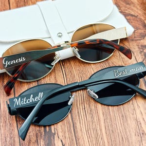 May include: Two pairs of sunglasses with personalized text. One pair has a gold frame with brown lenses and tortoise-shell arms, the other is black with dark lenses. The text on the frames reads "Genesis" and "Best man" and "Mitchell".