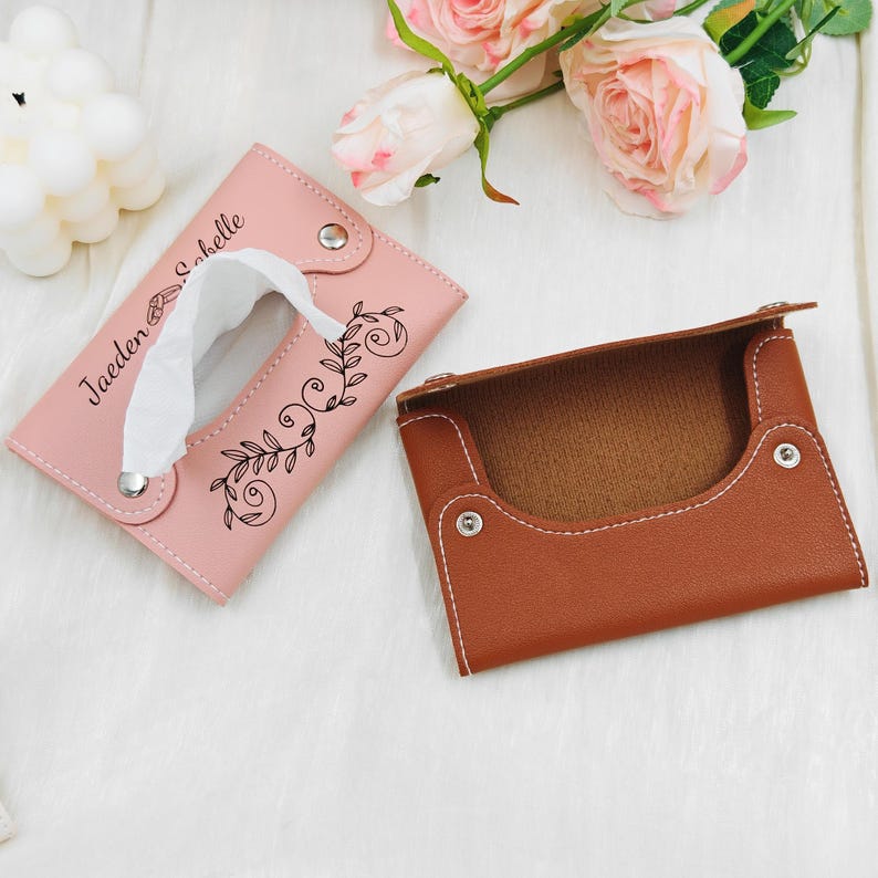 May include: A pink leather tissue box cover with a floral design and the name "Jaeden & Sabelle" printed on it. Next to it is a brown leather box with white stitching. The background includes pink roses and a white, fluffy surface.