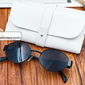 May include: Black-framed sunglasses with black mirror lenses rest on a wooden surface next to a white leather case. The sunglasses have an oval shape and a sleek, modern design. The case is closed with a strap and a silver button.