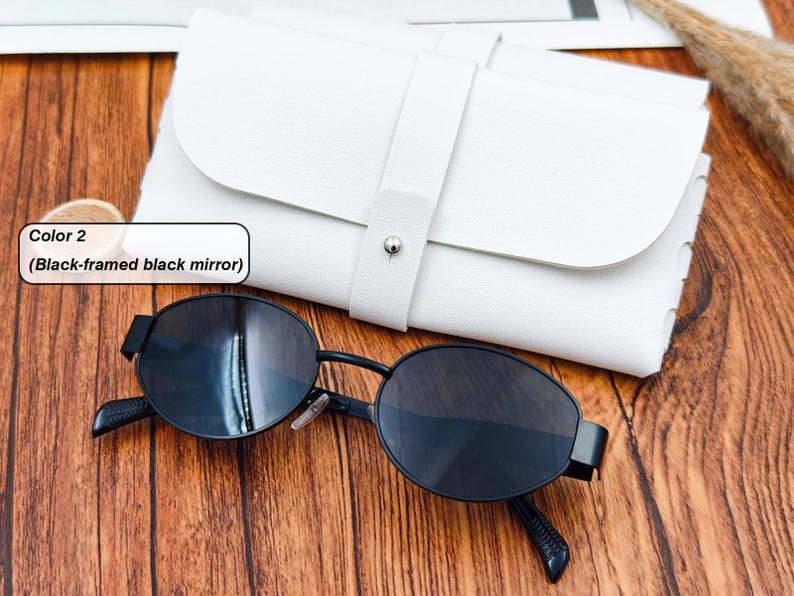 May include: Black-framed sunglasses with black mirror lenses rest on a wooden surface next to a white leather case. The sunglasses have an oval shape and a sleek, modern design. The case is closed with a strap and a silver button.