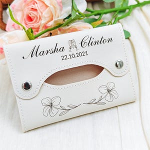 May include: Ivory-colored tissue holder with the names "Marsha" and "Clinton" and the date "22.10.2021" printed in black. The holder features a floral design and two silver snap closures. The background includes pink roses.