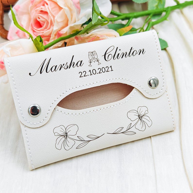 May include: Ivory-colored tissue holder with the names "Marsha" and "Clinton" and the date "22.10.2021" printed in black. The holder features a floral design and two silver snap closures. The background includes pink roses.