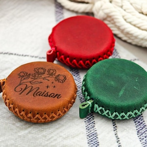 Può includere: Tre metri a nastro rotondi in pelle rossa, verde e marrone. Il metro a nastro marrone ha incisa la parola "Maison" e disegni floreali. Ogni metro a nastro ha un anello ed è cucito sui bordi.