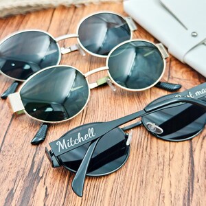 May include: Sunglasses with black lenses and silver and gold frames are arranged on a wooden surface. One pair has the name "Mitchell" printed on the arm. A white leather case is visible in the background.