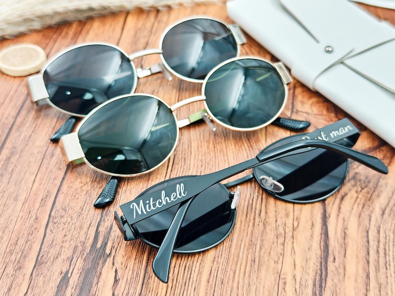 May include: Sunglasses with black lenses and silver and gold frames are arranged on a wooden surface. One pair has the name "Mitchell" printed on the arm. A white leather case is visible in the background.
