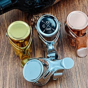 May include: A selection of wine bottle stoppers in gold, silver, and copper. One stopper features a black and silver design with the text "C R 2025". The stoppers are displayed on a wooden surface.