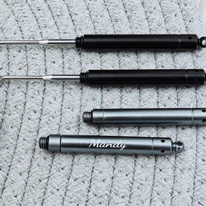 May include: An assortment of ear wax removal tools. Two black tools with silver tips and two silver tools, one engraved with the name "Mandy". The tools are on a gray woven surface.