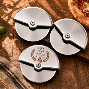Könnte beinhalten: Drei kreisförmige Pizzaschneider aus Edelstahl mit metallischem Finish. Einer der Schneider ist mit einem Lorbeerkranz, drei Sternen, einem Pizzastück-Symbol und dem Text Angela & Howard, Aug 2024 auf dem Griff graviert.