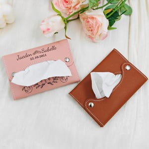 May include: Two leather tissue holders, one pink and one brown, are displayed. The pink holder has the names "Jaeden & Sabelle" and the date "22.10.2021" printed on it, along with a floral design. Both holders have white stitching and silver snap closures.