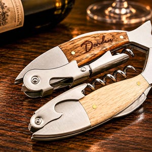 May include: Two fish-shaped corkscrews with metal components and wooden handles. The top corkscrew features a darker wood grain with the name Darden engraved in script. Both tools include a folding metal worm, a foil cutter, and a bottle opener.