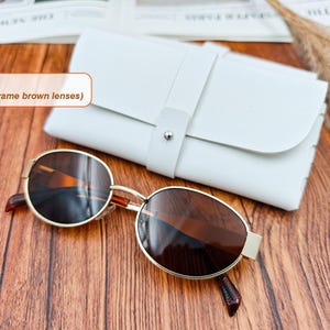 May include: Gold-framed sunglasses with brown lenses rest on a wooden surface next to a white leather case. The sunglasses have a classic oval shape. The case is closed with a strap and a silver button. The text in the image says "Color 1 (gold frame brown lenses)".