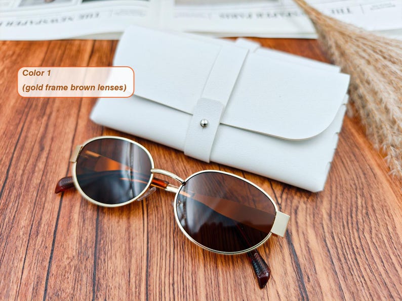May include: Gold-framed sunglasses with brown lenses rest on a wooden surface next to a white leather case. The sunglasses have a classic oval shape. The case is closed with a strap and a silver button. The text in the image says "Color 1 (gold frame brown lenses)".