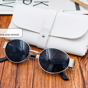 May include: Silver frame sunglasses with gray lenses and a white leather case. The sunglasses have a round shape and a silver frame. The case is rectangular with a strap closure. The text in the image says "Color 4 (Silver frame gray lenses)".