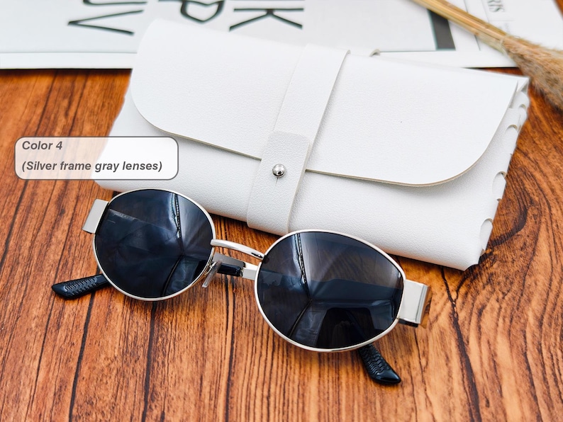May include: Silver frame sunglasses with gray lenses and a white leather case. The sunglasses have a round shape and a silver frame. The case is rectangular with a strap closure. The text in the image says "Color 4 (Silver frame gray lenses)".