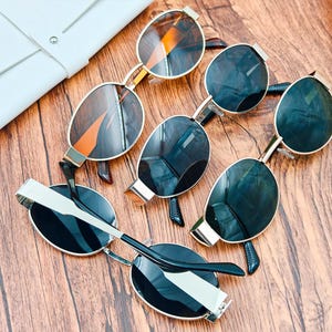 May include: An assortment of sunglasses with gold frames and dark lenses are arranged on a wooden surface. A white leather case is visible in the upper left corner. The sunglasses have a modern, stylish design.