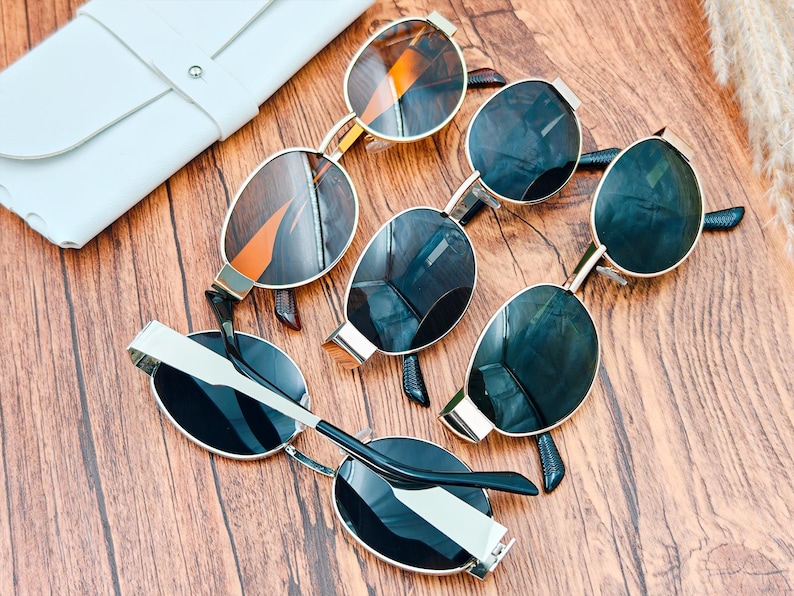 May include: An assortment of sunglasses with gold frames and dark lenses are arranged on a wooden surface. A white leather case is visible in the upper left corner. The sunglasses have a modern, stylish design.