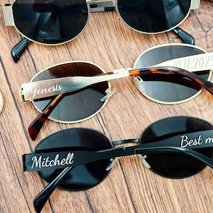 May include: Three pairs of sunglasses with dark lenses and personalized text on the frames. One pair is gold-toned, one has a tortoiseshell pattern, and the third is black. The text includes names and a date.