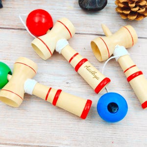 Puede incluir: Juguetes kendama de madera con detalles rojos y blancos y bolas de colores. Uno de los kendamas tiene grabado el nombre "Aadhya". Los juguetes están sobre una superficie de madera clara, con una bola roja, verde y azul.